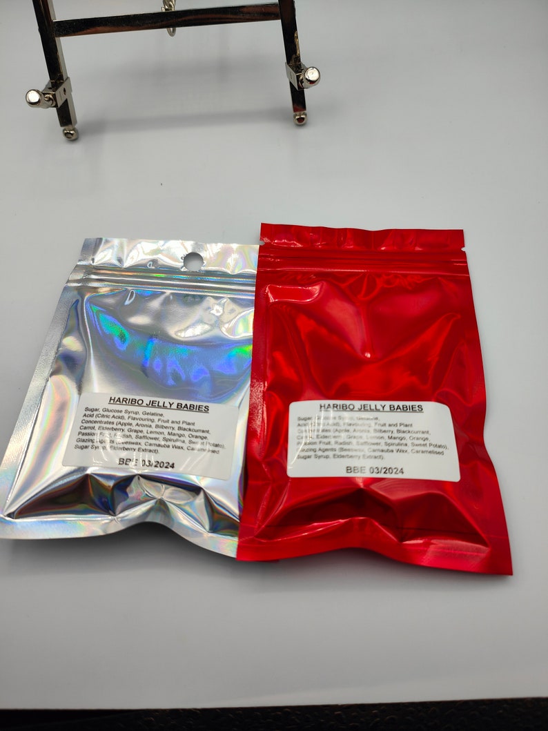 K&ouml;nnte beinhalten: Zwei rote und silberne Folienbeutel mit Haribo Jelly Babies S&uuml;&szlig;waren. Der rote Beutel hat ein Mindesthaltbarkeitsdatum vom 03/2024. Der silberne Beutel hat ein Mindesthaltbarkeitsdatum vom 13/2024. Die Beutel sind mit den Zutaten und N&auml;hrwertangaben gekennzeichnet.