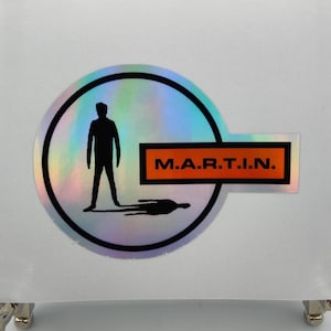 Könnte beinhalten: Holografischer Aufkleber mit einer schwarzen Silhouette einer Person in einem schwarz umrandeten Kreis. Der Aufkleber hat ein rechteckiges oranges Etikett mit dem weißen Text "M.A.R.T.I.N."