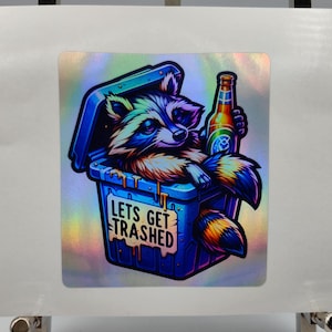 Könnte beinhalten: Holografischer Aufkleber mit einem Cartoon-Waschbären in einer blauen Mülltonne, der eine Bierflasche hält. Auf der Mülltonne steht der Text "LETS GET TRASHED". Der Waschbär hat farbenfrohes Fell und einen schelmischen Ausdruck.