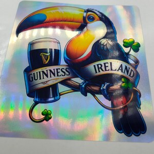 Op de afbeelding: Een toekan met een kleurrijke snavel en veren houdt een glas Guinness-bier vast. De toekan zit op een tak met een banner waarop "GUINNESS" en "IRLAND" staat. De achtergrond is een glinsterende, iriserende folie.