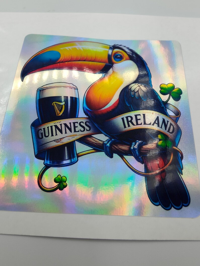 Op de afbeelding: Een toekan met een kleurrijke snavel en veren houdt een glas Guinness-bier vast. De toekan zit op een tak met een banner waarop "GUINNESS" en "IRLAND" staat. De achtergrond is een glinsterende, iriserende folie.