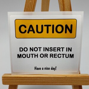 May include: A white sticker with a yellow rectangle that reads "CAUTION" in bold black letters. Below, it says "DO NOT INSERT IN MOUTH OR RECTUM" and "Have a nice day!" in black text.