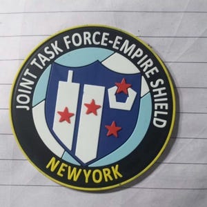 May include: A black and yellow circular patch with the text "Joint Task Force-Empire Shield" and "New York" in white. The patch features a blue and white shield with two white towers and three red stars.