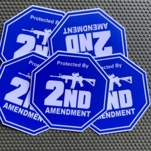 Könnte beinhalten: Ein Satz von vier blauen und weißen achteckigen Aufklebern mit dem Text "2nd Amendment" und einer Silhouette eines Gewehrs. Der Text "Protected By" befindet sich ebenfalls auf den Aufklebern.