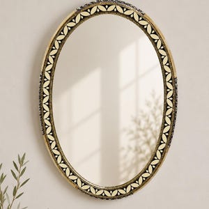 Può includere: Uno specchio da parete ovale con una cornice decorativa caratterizzata da un motivo a foglie ripetuto in nero e crema. La cornice presenta una finitura oro metallizzato con dettagli testurizzati sul bordo esterno, attorno a una superficie in vetro.