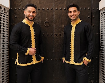 May include: Two black shirts with gold trim and accents. The shirts have long sleeves and a mandarin collar. The gold trim is along the front, collar, and cuffs. The shirts are paired with black pants. The background is a dark wooden door.