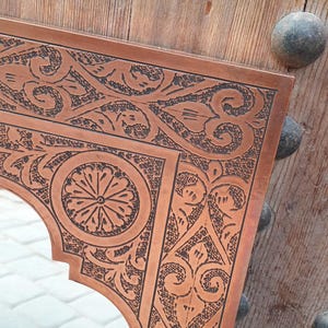 May include: Ornate copper mirror with intricate floral and geometric patterns. The mirror's frame has a warm, reddish-brown hue and is set against a wooden door with decorative metal studs. The design is detailed and eye-catching.