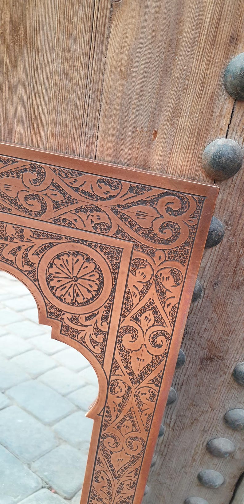 May include: Ornate copper mirror with intricate floral and geometric patterns. The mirror's frame has a warm, reddish-brown hue and is set against a wooden door with decorative metal studs. The design is detailed and eye-catching.