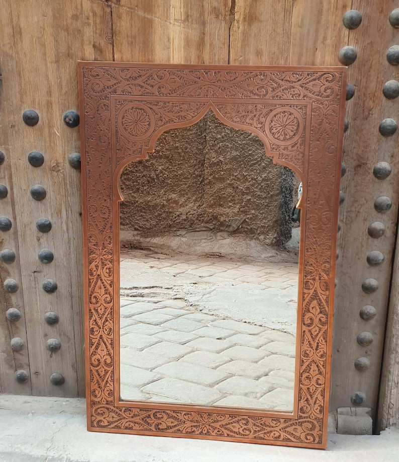 May include: A rectangular, brown, ornate mirror with an arched top. The frame features intricate floral and geometric carvings. The mirror reflects a stone-paved area. The mirror is leaning against a wooden door with metal studs.