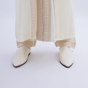 May include: A pair of white leather slippers with pointed toes. The slippers have a simple design and are worn with a white robe with gold embroidery.