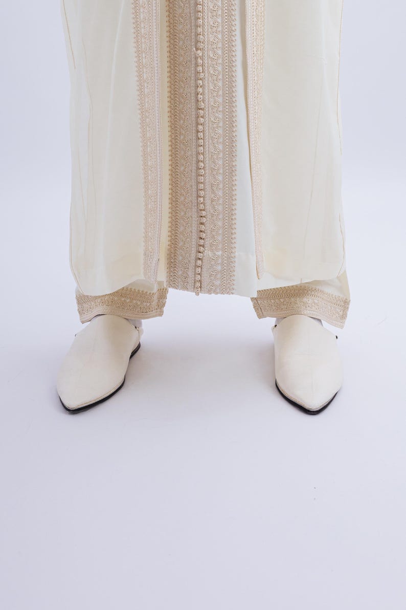 May include: A pair of white leather slippers with pointed toes. The slippers have a simple design and are worn with a white robe with gold embroidery.