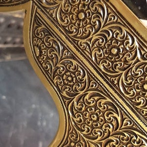 Pode incluir: Close-up de uma moldura de metal dourada com intrincados desenhos florais. A moldura tem uma superf&iacute;cie texturizada em relevo e uma forma ligeiramente curva.