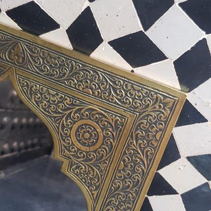 K&ouml;nnte beinhalten: Ein goldfarbener Metallrahmen mit aufwendigen floralen Designs. Der Rahmen ist vor einem Hintergrund aus schwarzen und wei&szlig;en rautenf&ouml;rmigen Fliesen platziert.