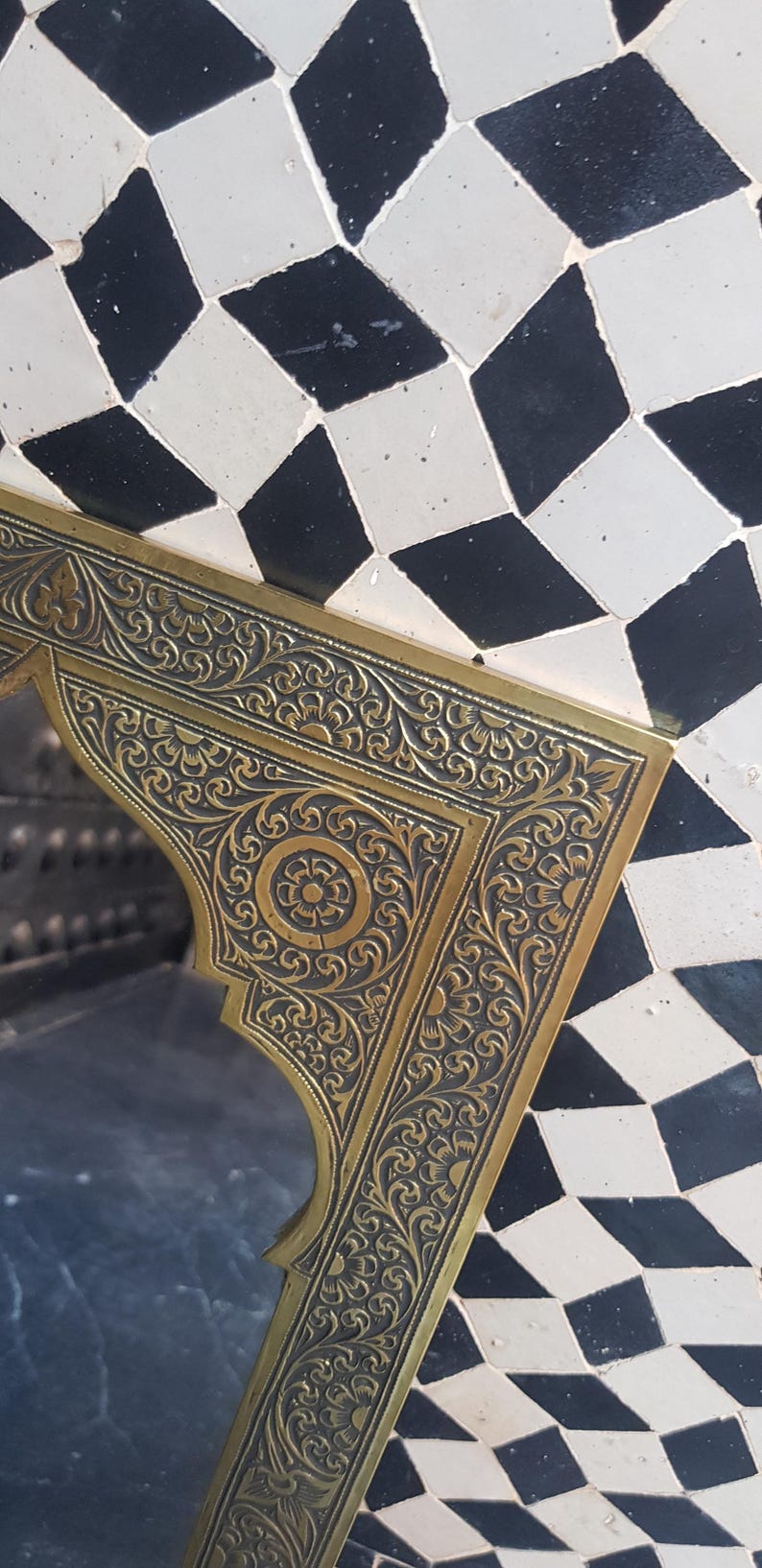 K&ouml;nnte beinhalten: Ein goldfarbener Metallrahmen mit aufwendigen floralen Designs. Der Rahmen ist vor einem Hintergrund aus schwarzen und wei&szlig;en rautenf&ouml;rmigen Fliesen platziert.