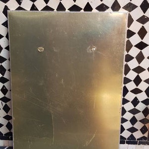Pode incluir: Uma placa de metal dourada com dois pequenos ganchos redondos de metal na parte superior. A placa est&aacute; pendurada em uma parede com um padr&atilde;o geom&eacute;trico preto e branco.