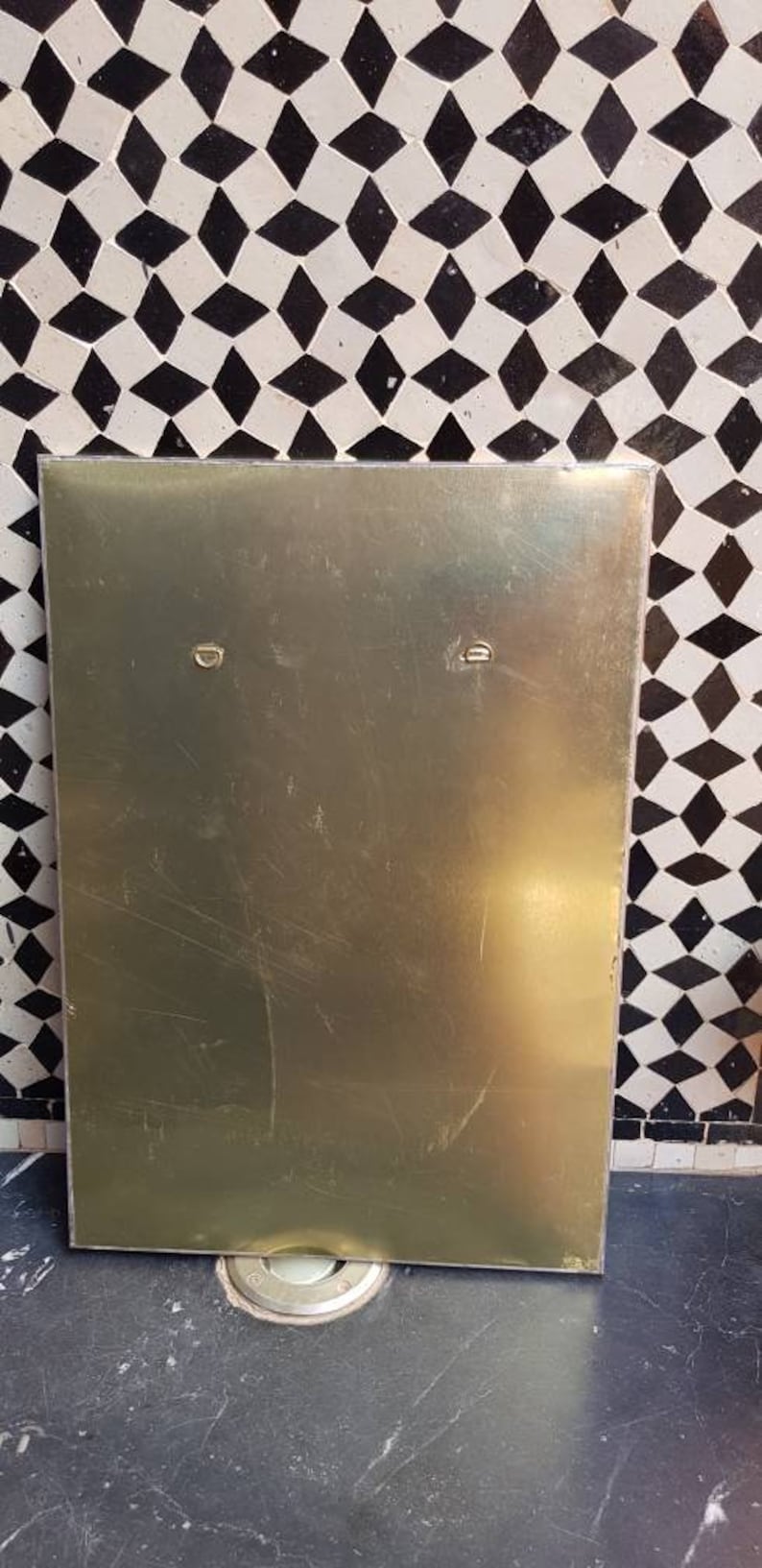 Pode incluir: Uma placa de metal dourada com dois pequenos ganchos redondos de metal na parte superior. A placa est&aacute; pendurada em uma parede com um padr&atilde;o geom&eacute;trico preto e branco.