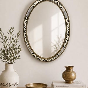 Può includere: Uno specchio da parete ovale con una cornice metallica scura e testurizzata, caratterizzata da un motivo ripetitivo di foglie color crema e accenti neri. Lo specchio riflette la luce e presenta un bordo decorativo con motivi floreali e geometrici.