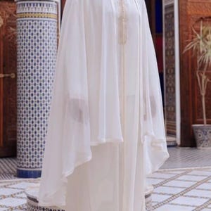 May include: A cream-colored traditional Moroccan garment, featuring a long robe and a sheer cape. The robe has gold embroidery around the neckline. The person is wearing white socks and cream-colored shoes. The background includes ornate architecture.