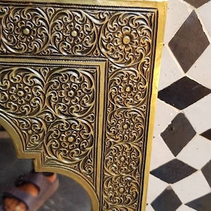 Pode incluir: Um espelho adornado de cor dourada com uma moldura decorativa que apresenta padr&otilde;es florais intrincados. O espelho &eacute; retangular com uma parte superior arqueada e est&aacute; colocado sobre um fundo de azulejos pretos e brancos.