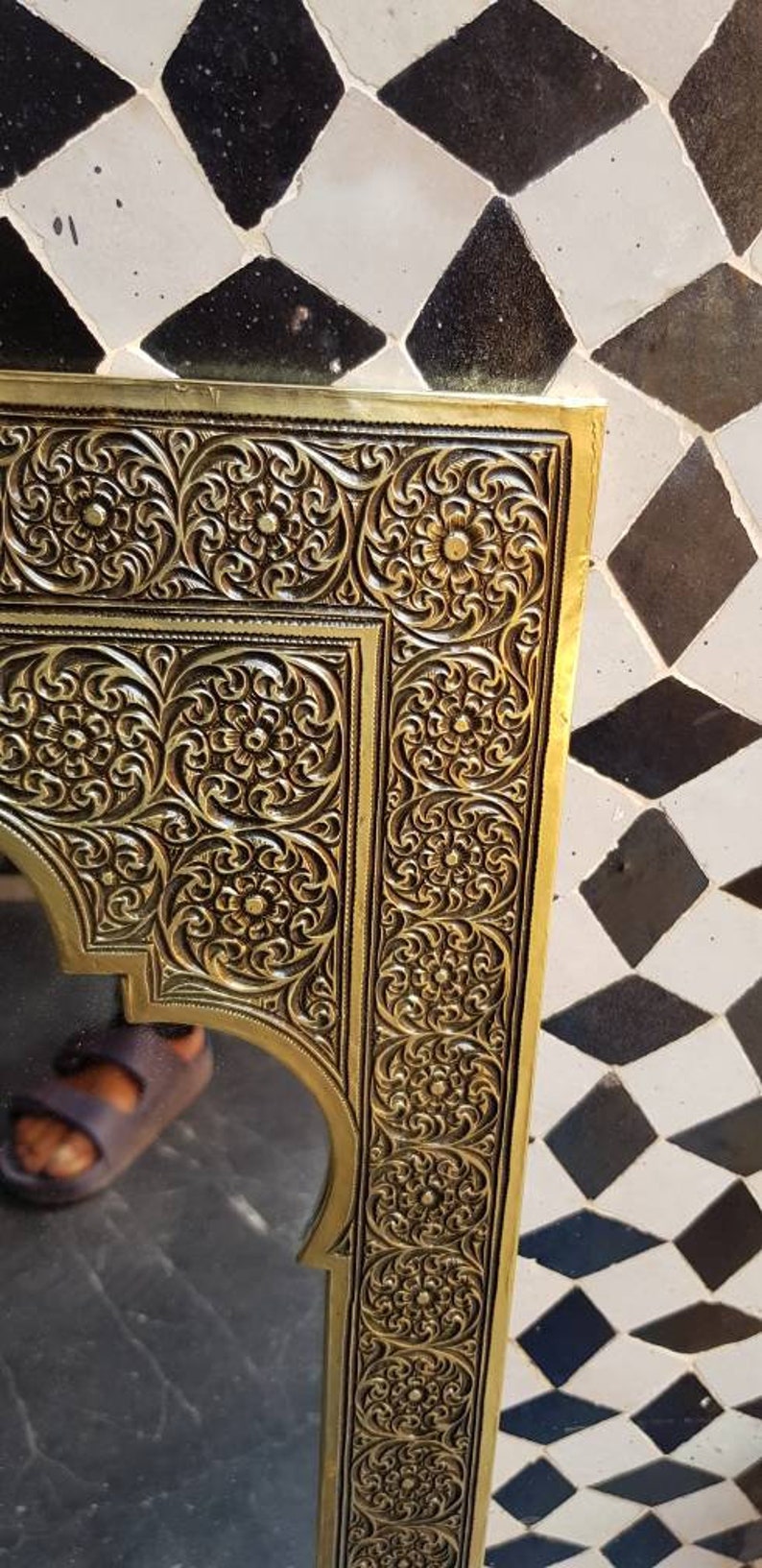 Pode incluir: Um espelho adornado de cor dourada com uma moldura decorativa que apresenta padr&otilde;es florais intrincados. O espelho &eacute; retangular com uma parte superior arqueada e est&aacute; colocado sobre um fundo de azulejos pretos e brancos.