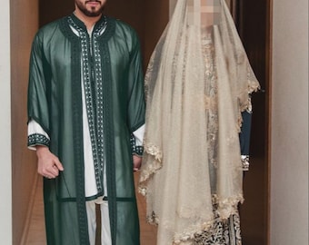 May include: A couple in traditional Moroccan wedding attire. The man is wearing a green robe with white embroidery and yellow slippers. The woman is wearing a long green dress with gold embroidery and a white veil.