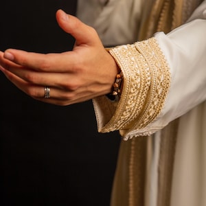 May include: A close-up of a white garment with gold embroidery. The garment has a long sleeve with a decorative cuff. The hand of the person wearing the garment is outstretched with a ring on the finger.