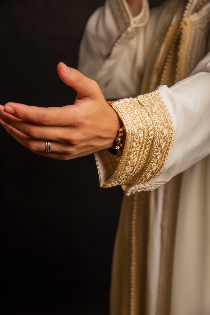 May include: A close-up of a white garment with gold embroidery. The garment has a long sleeve with a decorative cuff. The hand of the person wearing the garment is outstretched with a ring on the finger.