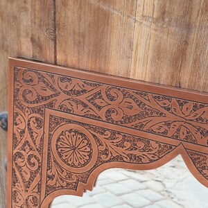 May include: Close-up of a brown wooden mirror frame with intricate, dark etched floral and geometric patterns. The frame has an arched bottom edge and is set against a wooden background.