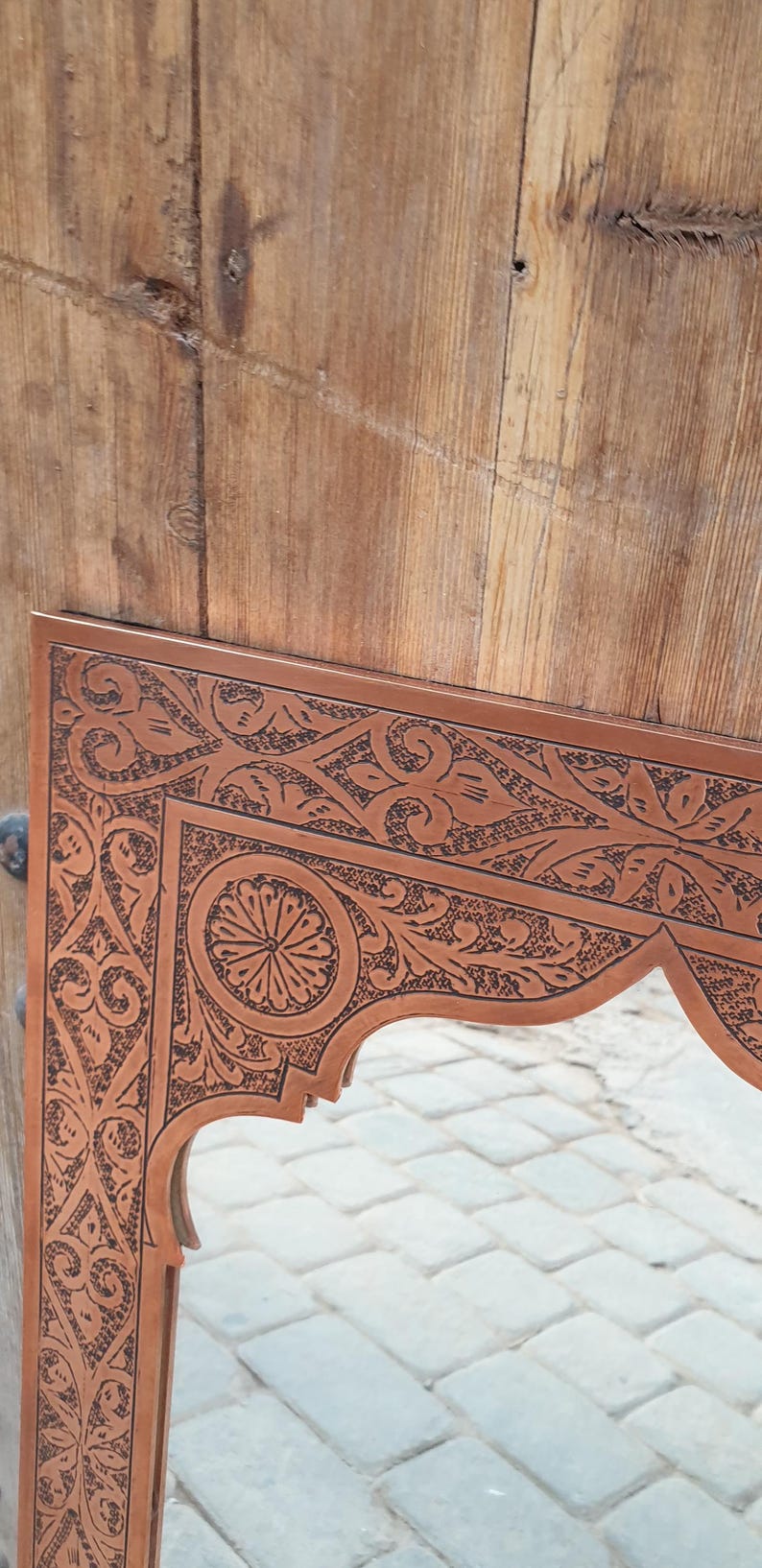 May include: Close-up of a brown wooden mirror frame with intricate, dark etched floral and geometric patterns. The frame has an arched bottom edge and is set against a wooden background.