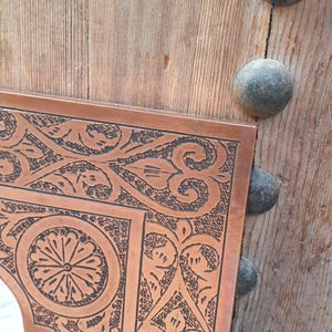 May include: Close-up of a copper-colored mirror frame with intricate black etched floral and geometric designs. The frame is set against a weathered wooden door with dark metal studs.