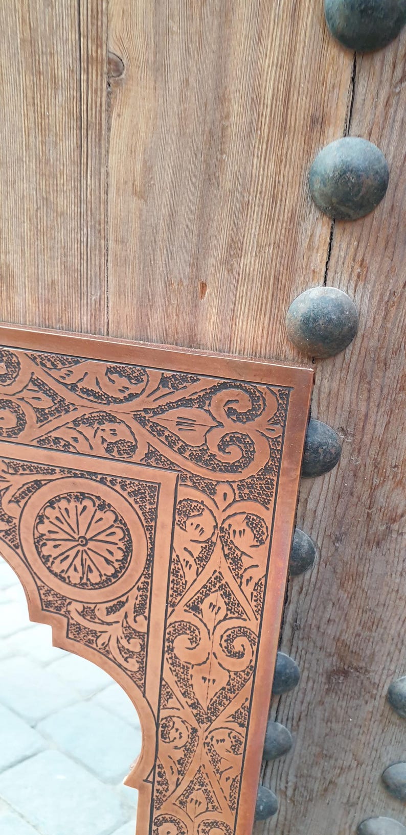 May include: Close-up of a copper-colored mirror frame with intricate black etched floral and geometric designs. The frame is set against a weathered wooden door with dark metal studs.