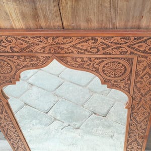 May include: A rectangular, ornate wooden mirror with a carved floral and geometric design. The mirror has an arched top and is set against a wooden background. The frame is a warm brown color.