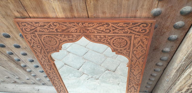 May include: A rectangular, ornate wooden mirror with a carved floral and geometric design. The mirror has an arched top and is set against a wooden background. The frame is a warm brown color.