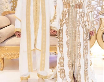 Könnte beinhalten: Ein Paar in traditioneller weißer und goldfarbener bestickter Kleidung. Das Kleid der Frau hat einen langen, fließenden Rock und ein tailliertes Oberteil. Das Outfit des Mannes beinhaltet eine lange Robe mit goldenen Verzierungen.