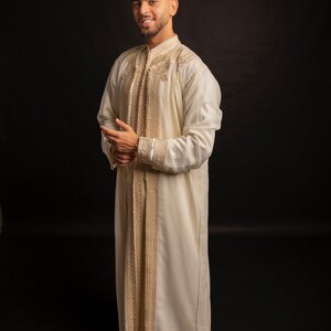 May include: A man wearing a white robe with gold embroidery and yellow slippers. The robe has a long, flowing design and is decorated with intricate patterns.