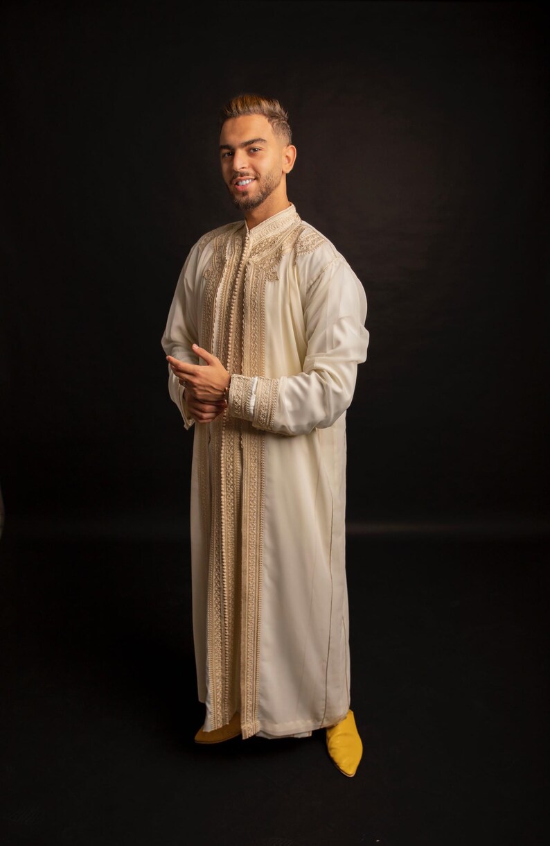 May include: A man wearing a white robe with gold embroidery and yellow slippers. The robe has a long, flowing design and is decorated with intricate patterns.