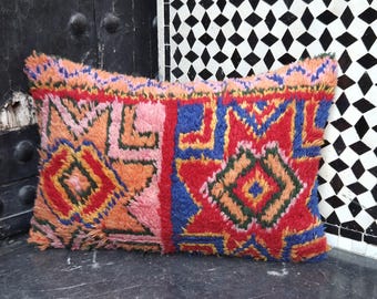 May include: Rectangular, colorful pillow with a geometric pattern. The pillow features a mix of red, blue, orange, pink, and yellow hues. The design includes diamond and star-like shapes. The pillow is made of a textured material.