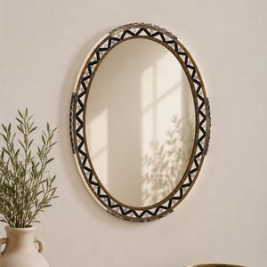 Può includere: Uno specchio da parete ovale con una cornice decorativa caratterizzata da un motivo a zigzag bianco e nero. Lo specchio presenta una superficie in vetro riflettente e un bordo esterno scuro e testurizzato. Il design include dettagli complessi.
