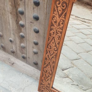 May include: A rectangular brown mirror with an ornate, carved frame featuring a repeating floral and geometric pattern. The mirror is leaning against a wooden door with dark, rounded studs.