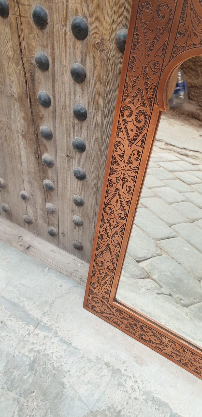 May include: A rectangular brown mirror with an ornate, carved frame featuring a repeating floral and geometric pattern. The mirror is leaning against a wooden door with dark, rounded studs.