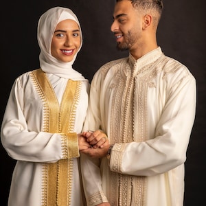 May include: A couple wearing traditional white clothing with gold embroidery. The woman is wearing a headscarf. They are holding hands and smiling at each other.
