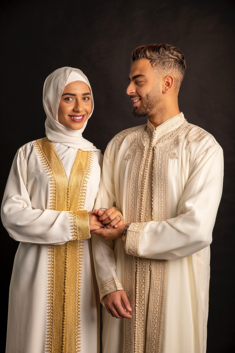 May include: A couple wearing traditional white clothing with gold embroidery. The woman is wearing a headscarf. They are holding hands and smiling at each other.