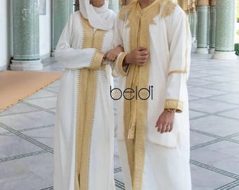 May include: A couple wearing traditional white clothing with gold embroidery. The man is wearing a white robe with gold trim and the woman is wearing a long white dress with gold embroidery. They are standing in front of a building with columns.