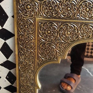 Pode incluir: Um espelho de metal dourado com um design ornamentado, intrincado e detalhado. O espelho tem uma forma de arco marroquino tradicional e &eacute; emoldurado por uma moldura decorativa.