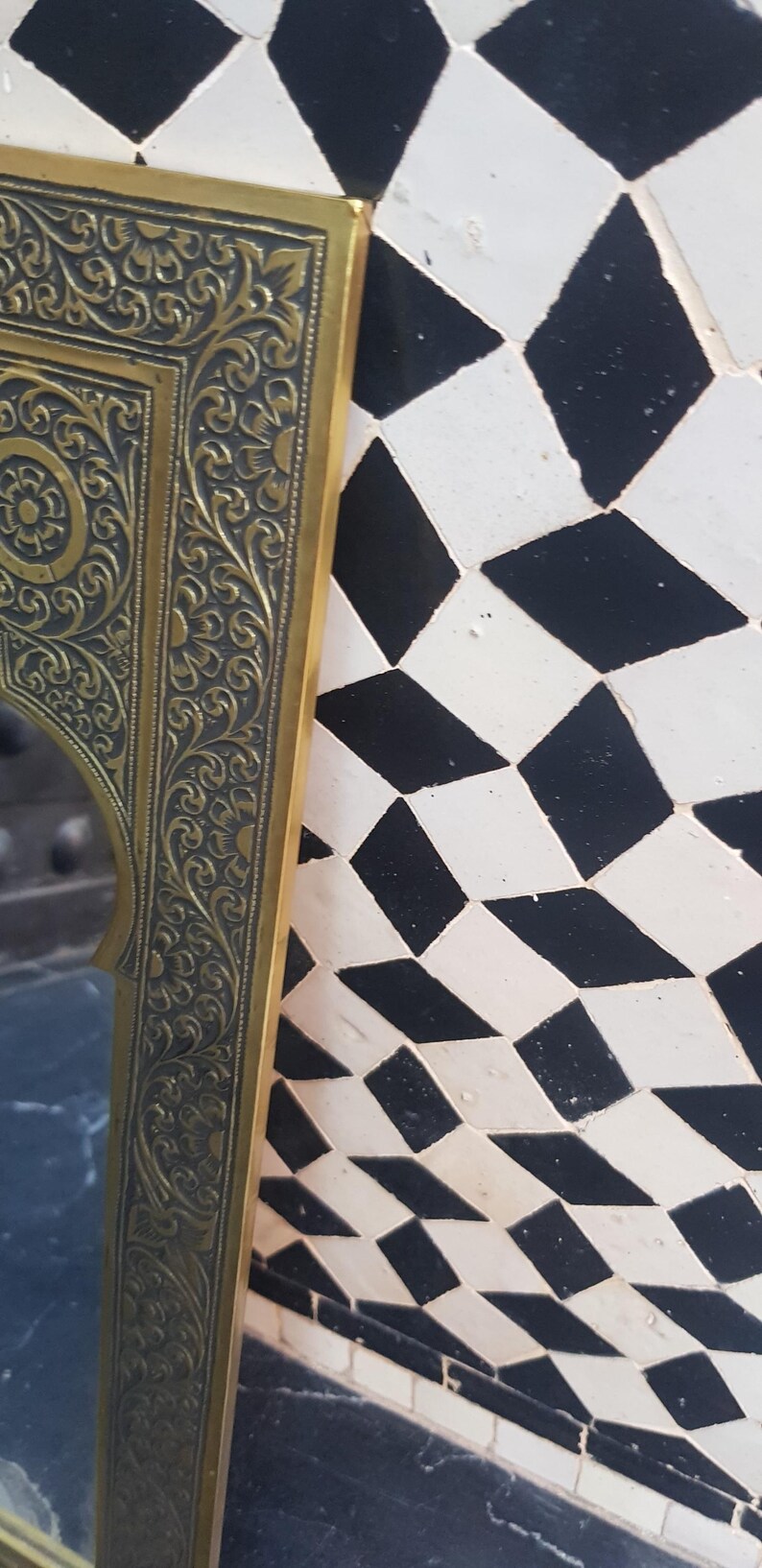 K&ouml;nnte beinhalten: Ein verzierter Spiegelrahmen in Gold mit aufwendigen floralen Mustern. Der Rahmen ist vor einem Hintergrund aus schwarzen und wei&szlig;en Diamantenfliesen platziert.