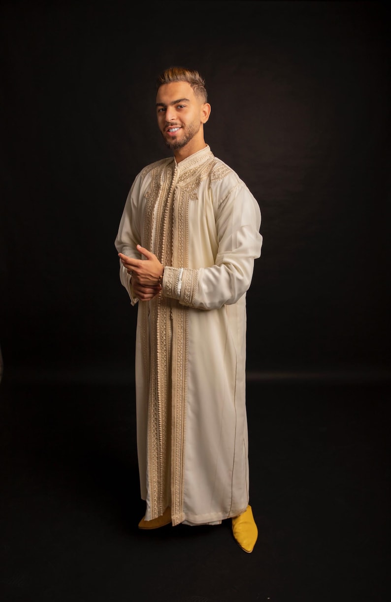 May include: A man wearing a white robe with gold embroidery and yellow slippers. The robe has a long, flowing design and is decorated with intricate patterns.
