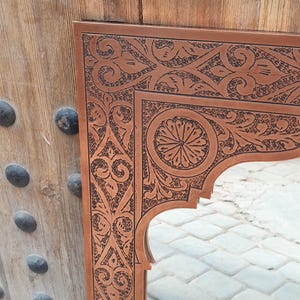 May include: A brown wooden mirror frame with intricate, dark brown carved floral and geometric patterns. The frame has an arched top and is set against a wooden door with metal studs.