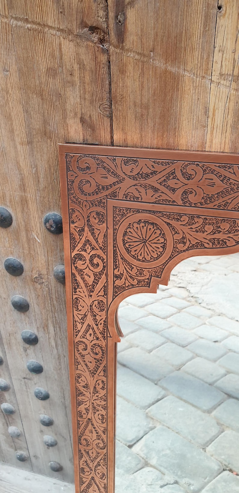 May include: A brown wooden mirror frame with intricate, dark brown carved floral and geometric patterns. The frame has an arched top and is set against a wooden door with metal studs.