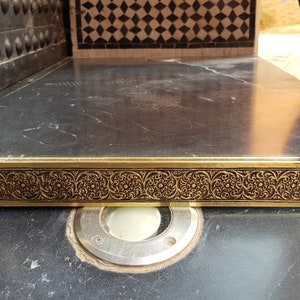 Pode incluir: Um espelho adornado de cor dourada com um design detalhado e intrincado em torno da moldura. O espelho reflete um piso de ladrilhos.