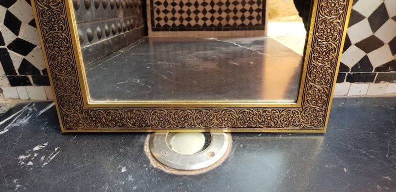 Pode incluir: Um espelho adornado de cor dourada com um design detalhado e intrincado em torno da moldura. O espelho reflete um piso de ladrilhos.